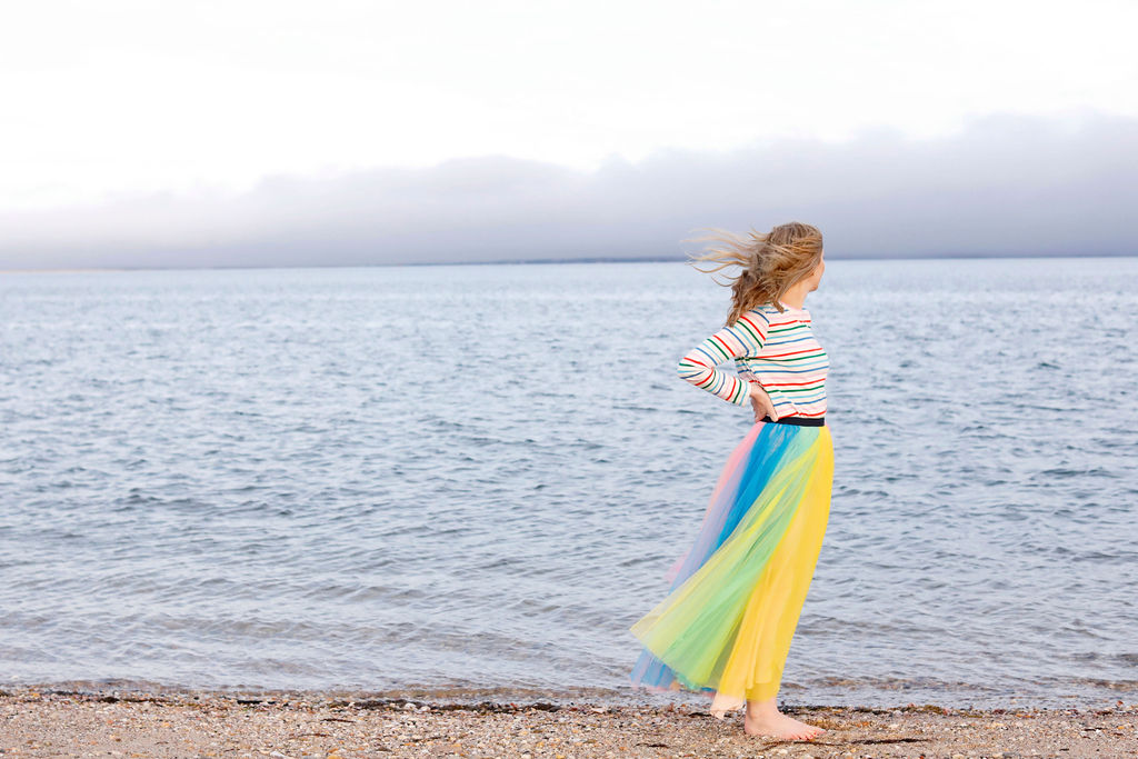 An image of a woman in a striped top and multicolored skirt standing on a beach looking out at the ocean. 