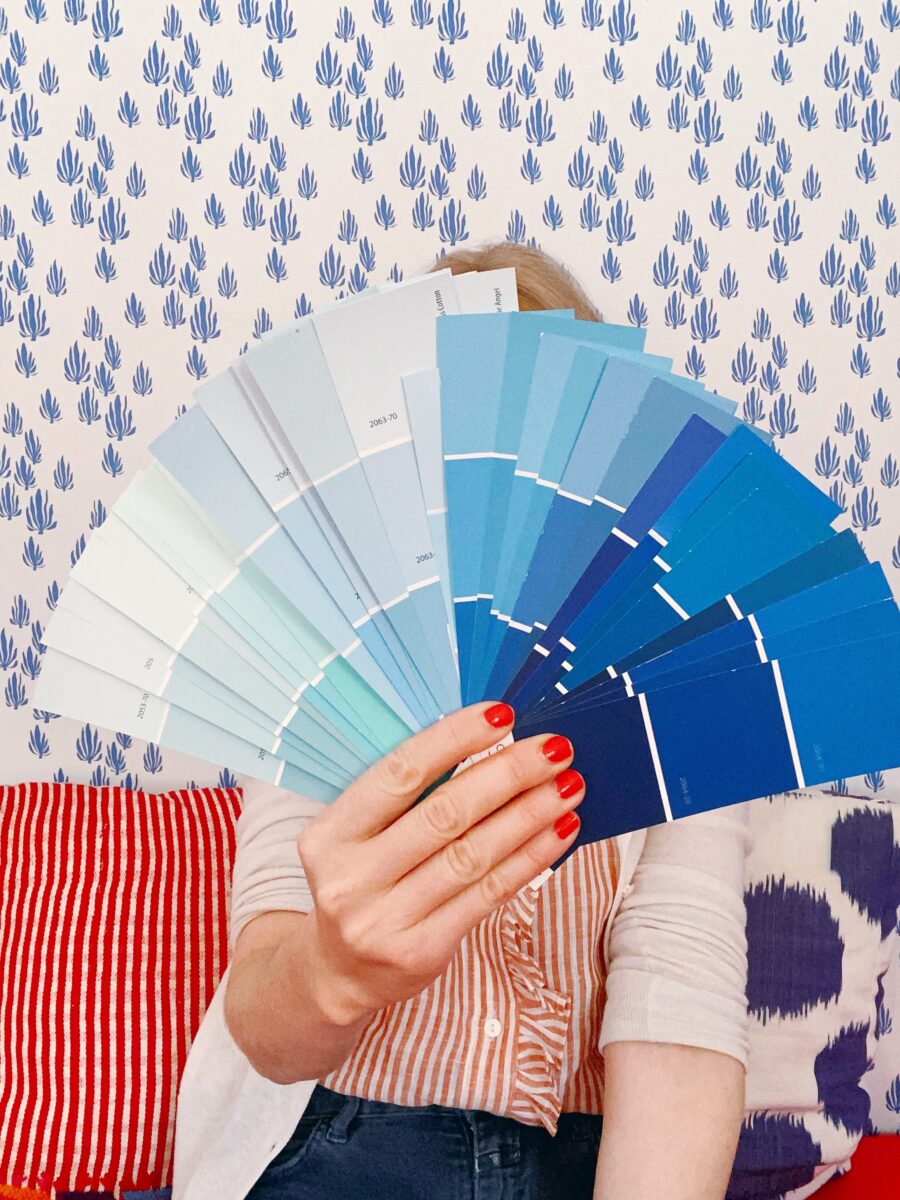 Ingrid holding an array of blue paint swatches against a background of blue and white printed wallpaper