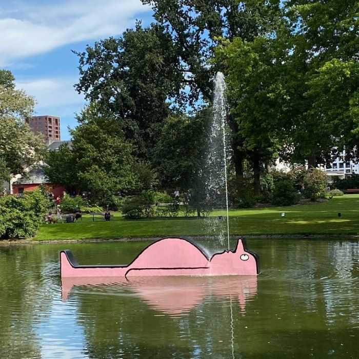 An installation of a cartoon character with a round belly lying on his back in a pond, with a fountain spraying water as if it is coming out of his mouth. 
