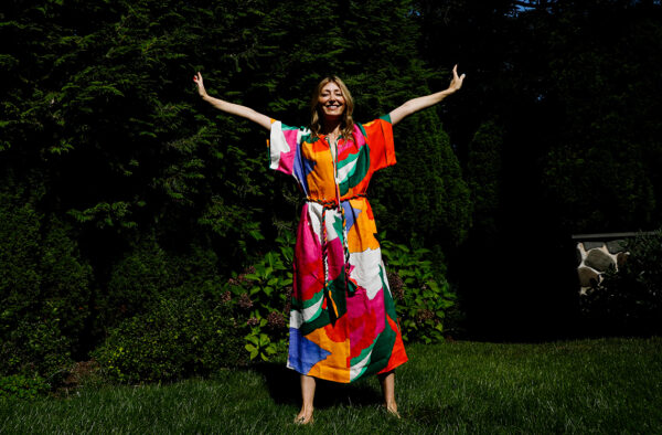 Image of Ingrid standing arms akimbo in a colorful abstract print dress surrounded by greenery.