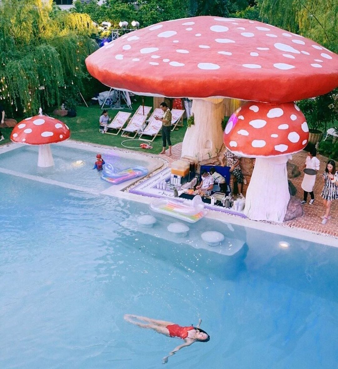 A swimming pool with giant red and white mushroom sculptures towering over it. 