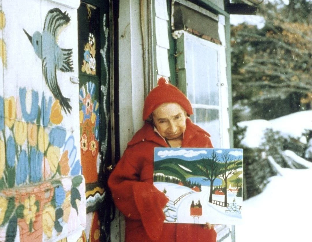 A picture of artist Maud Lewis wearing a red coat holding a winter landscape in front of her house in winter.