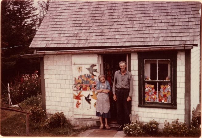 Maud and Everett Lewis in front of their home, featuring paintings on their doors and windows. 