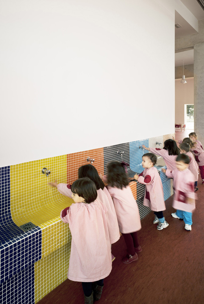 Young children in smocks wash hands at a long trough-style sink tiled in small tiles with large blocks of color. 