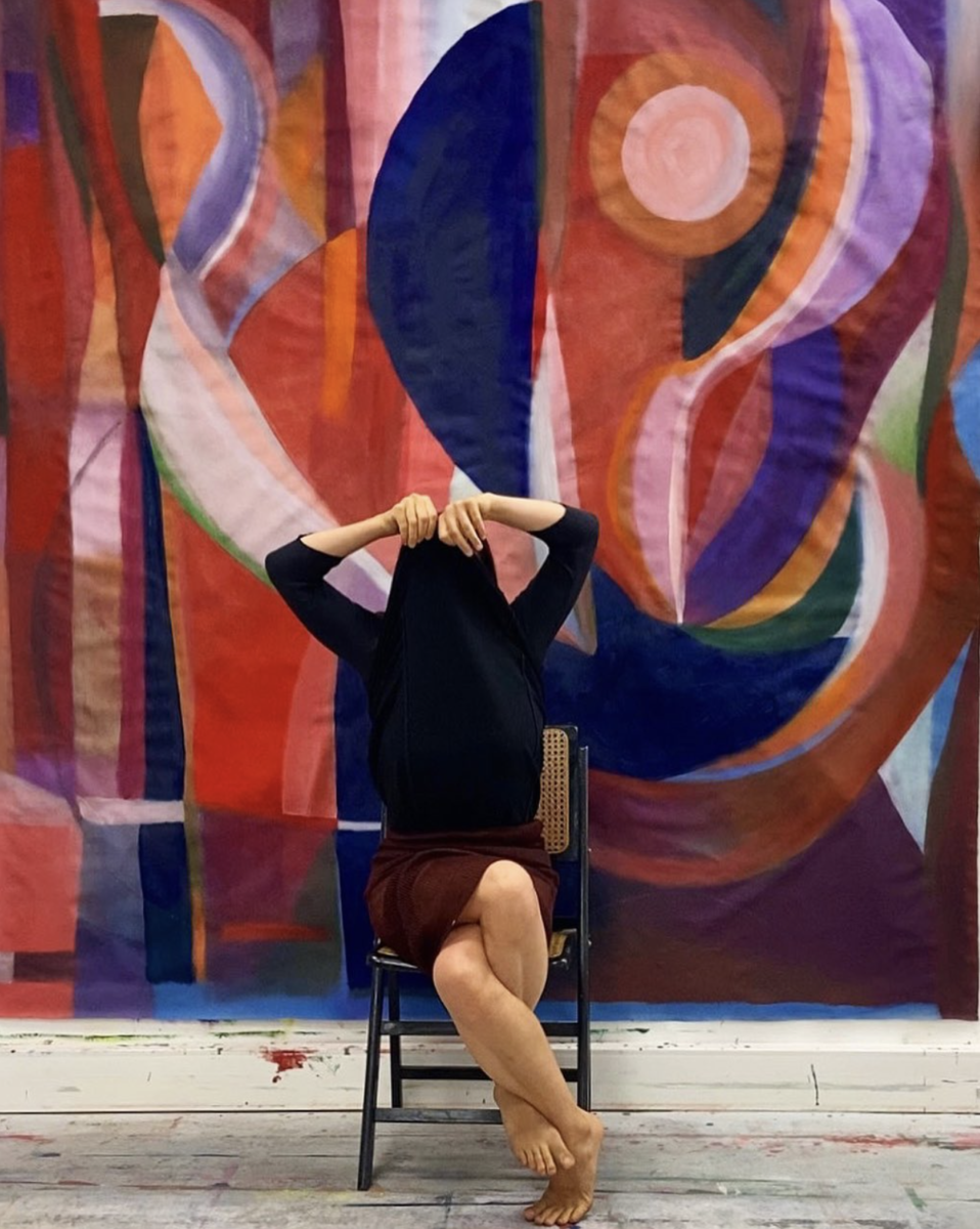 artist Caroline Denervaud sitting in a chair wearing black covering her face in front of a wall-sized painting of abstract shapes in terracotta orange, deep blue, lilac, and green. 