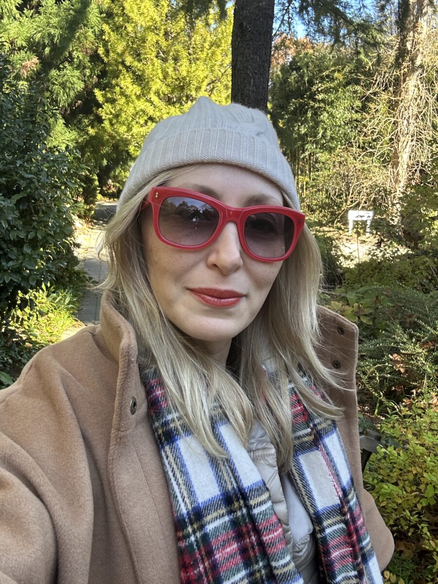 Image of Ingrid standing outside among trees wearing red sunglasses and a red lip gloss. 
