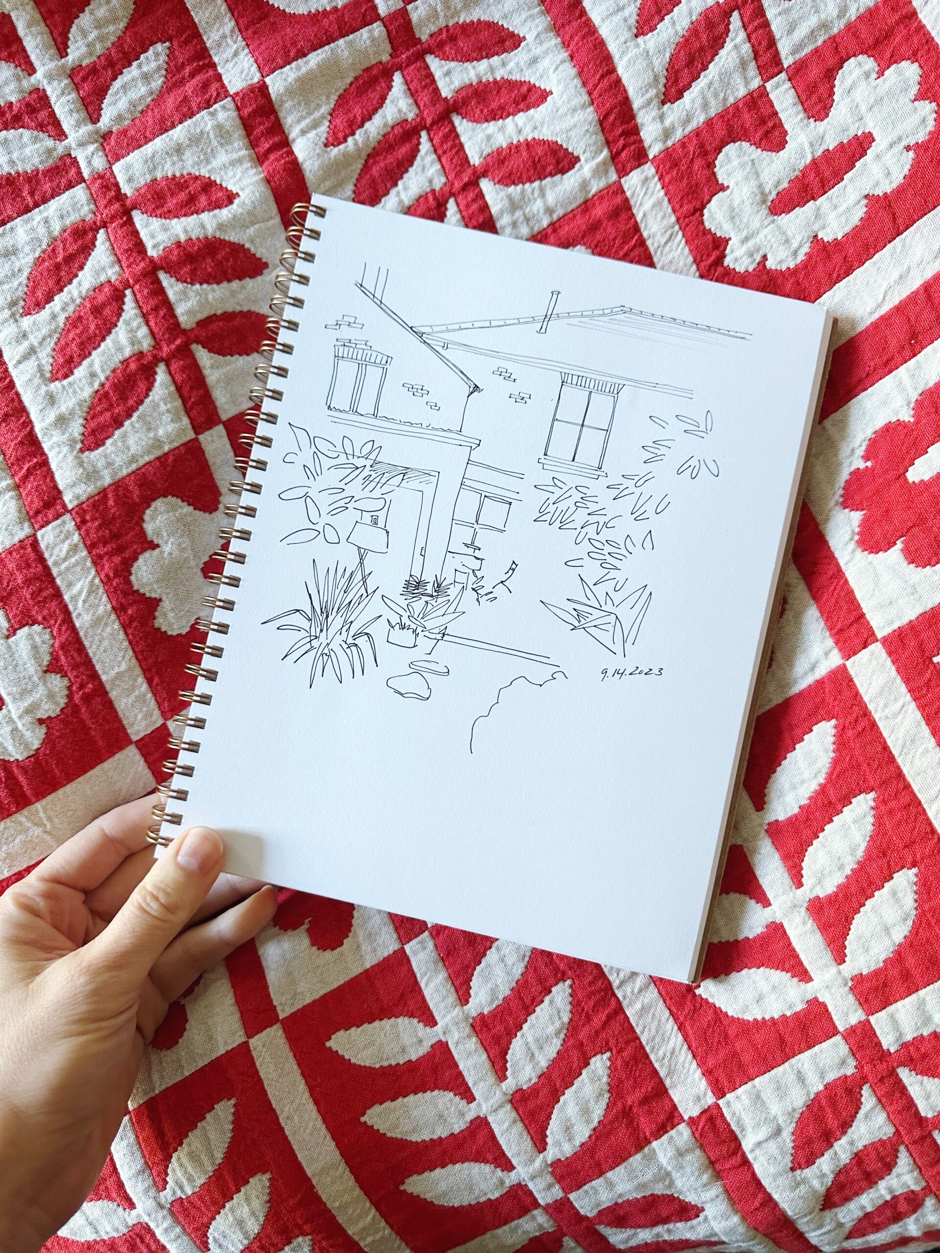 A drawing of a house and garden in ink in a notebook on a red and white leaf patterned quilt.