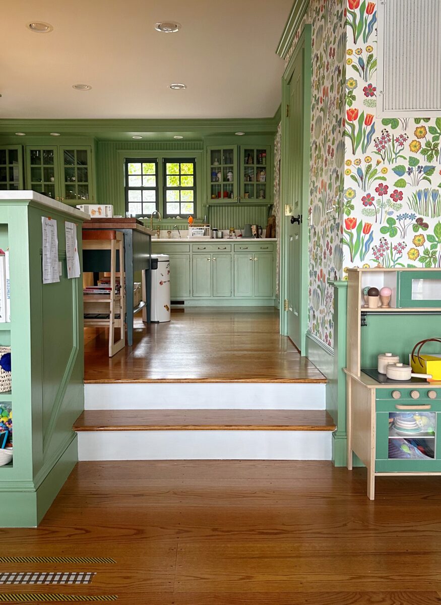 A view from a dining room into a kitchen painted green, with floral wallpaper and wood floors.